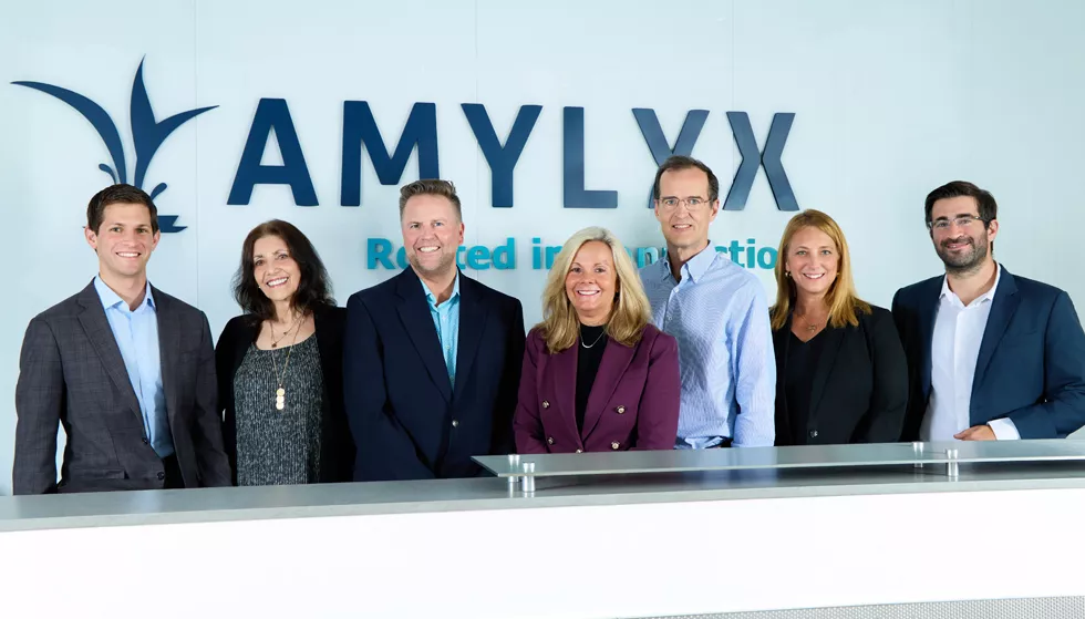 Amylyx leadership team smiling in front of the Amylyx sign. The sign reads, "Rooted in Connection."